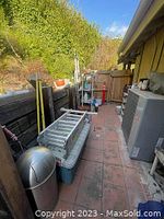 Wide-angle photo showing a large cooler with a folded aluminum ladder on top, several yellow-handled garden tools leaning against the wooden fence, a metallic outdoor waste bin, and other assorted outdoor items along a narrow paved walkway next to a building.