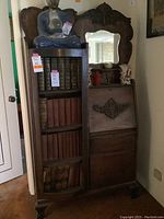 Full view of antique wooden cabinet showing carved top, bookshelf with vintage books, and decorative items on top including blue Buddha statue and porcelain figurines.