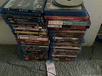 Stack of Blu-ray and DVD movie cases in two piles, showing titles such as The Wave, Jeepers Creepers, Poseidon, Stargate, The Woman King, Godzilla, Tremors, Jurassic World, Predator 2, and more.