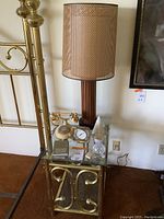Full view of brass and glass table holding wood lamp, rotary phone, clock, and decor items beside a brass bed frame.