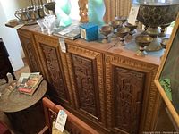 Front and top view of carved wooden drinks bar showing intricate carved panels and decorative geometric patterning on edges. Several metal and glass items on top for staging only.
