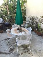 Full view of white metal patio dining table with green umbrella and four chairs on concrete surface outdoors with surrounding plants.