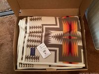 Opened cardboard box showing folded wool blanket with southwestern geometric design in beige, gray, red, orange, and brown.