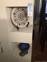 Overview of the inlaid stone plate displayed on a black stand and the cobalt blue glass vase with a metal base below it in a recessed shelf.