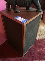 Full side view of one wooden CPI speaker resting on red carpet, showing wood grain finish and speaker grille.
