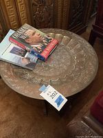 Photo of round metal table with detailed engraving and three books on top, showing table surface and part of the base.