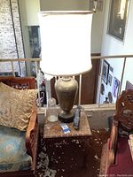 Metal lamp with floral engraved vase body and large white shade on small wood table with inlaid diamond pattern, visible oil lamp and golf statue on table.