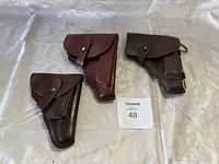 Three leather holsters of different shades and designs arranged on a white cloth surface.