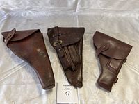 Three brown leather military holsters of various sizes and closures arranged side by side on a white cloth background.