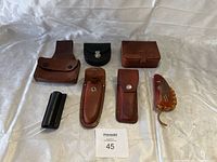 Top-down view of the assortment of 8 leather and synthetic holsters and pouches arranged on white fabric background showing various sizes and styles.