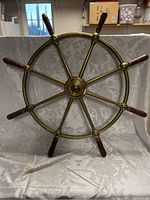 Full view of the 28 inch brass ship's wheel with eight spokes and wooden handles against a fabric backdrop.
