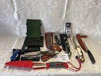 Photo showing the entire lot with knives, multi-tools, sheaths and green display box on a white patterned cloth