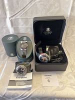 Three Citizen Eco-Drive watches displayed: pilot chronograph in green oval box with hang tag, dive-style watch on foam with manual, resin analog watch in black zippered box