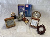 Full view of lot with eight clocks and timepieces arranged on fabric background showing variety of styles and cases.