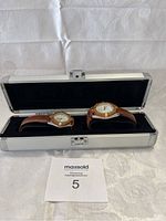 Pair of Crowley wristwatches with brown leather straps displayed inside open silver metal case with black velvet lining on white textured surface.