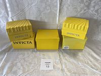 Five yellow Invicta watch boxes stacked and closed on a white cloth background.