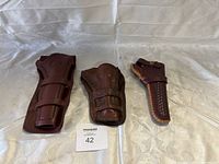 Lot of 3 leather holsters on a fabric surface: one dark brown large holster, one medium brown worn holster, one small light brown holster with detailed stitching.