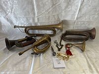 Seven brass musical instruments including various bugles and one horn with tassel on white fabric background.