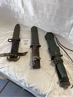 Three military knives with sheaths placed on a white surface, showing full length and handles.