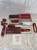 Seven Victorinox Swiss Army knives in red handles arranged on white background, with original boxes and a leather sheath visible.