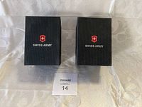 Two closed black Swiss Army watch boxes on cream fabric background.