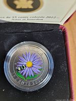 Clear close-up photo of the coin inside a protective case showing the colored aster flower with bumble bee and inscriptions.