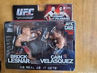 Front view of UFC 121 Brock Lesnar vs Cain Velasquez packaged action figures, showing both figures side by side in fighting poses with branding and display stand info visible.