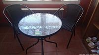 Two brown wicker chairs around round glass top table on tiled floor by window