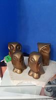 Four bronze owl figurines positioned on a white irregular-shaped stone base against a blue backdrop, showing the front and angled views with owl facial details.