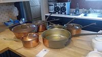 Seven pieces of French copper cookware including pots and pans with lids and long handles on a kitchen counter