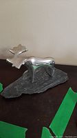 Front angle view of stylized polished silver metal moose sculpture mounted on rough dark stone base, showing signature on base.