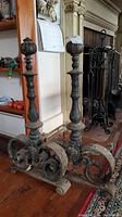 Pair of heavy metal andirons with decorative scroll bases and brass finial tops placed on wooden floor near fireplace.