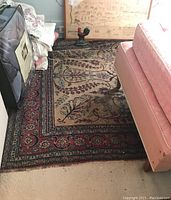 Photo showing the area rug beneath furniture including a pink upholstered seat and near framed artwork, displaying a floral and geometric design with darker border.