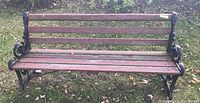 Photo of full front view of wrought iron and wood bench showing five wood slats on the seat and backrest with black decorative wrought iron ends and legs, bench placed on grass outdoors.