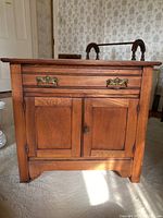 Front view of wooden dresser / wash stand showing drawer with ornate brass handles and two cabinet doors below.
