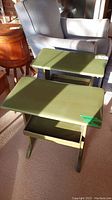Two olive green painted wood end tables with rectangular tops and open storage compartments underneath. Tables have curved legs and visible scratches.