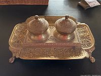 Front and top view of ornate metal double inkwell set on tray with decorative handles and claw feet.