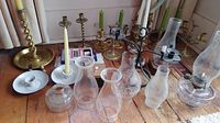 Group photo showing glass oil lamps, wood hurricane lamp, multiple brass candle holders, glass chimneys, and candles arranged on a wooden surface.