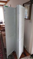 View of white wood three-panel privacy screen in folded position in a room next to furniture and window.
