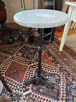 Full view of the stand showing the round stone top with a speckled grey and white pattern and black cast iron pedestal and base on patterned rug.