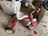 Left-side view of red and white tricycle showing frame, seat, wheels and pedals on garage floor