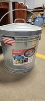 Photo showing the full 10 gallon metal trash can with locking lid and attached handle. Behrens branding and American flag label visible on the lid.