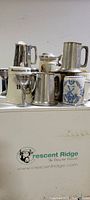 Front view of various size English pewter mugs and cups with handles, including a ceramic mug with blue windmill design and a pewter tankard with lid.