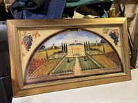 Full view of the framed painting depicting the Bordeaux wine region vineyard and chateau with grapevine accents in a gold painted cylindrical braided frame.