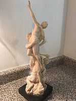 Side angle view of the white figurative statue showing two men and a woman with the woman raised high, mounted on a black base.