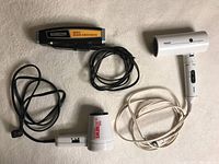 Three vintage travel-size hair drying and grooming devices displayed on a light carpet: one white Philips hair dryer with foldable handle, one small white HOT STYLER hair dryer with brush, and one black Salon Care mist hair groomer.