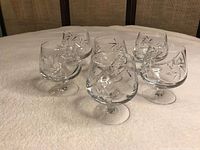 Six clear brandy snifter glasses arranged in a circle on a beige textured fabric surface, showing etched star patterns on bowls.