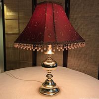 Full view of brass table lamp with lit bulb showing red fabric lampshade decorated with gold swirl patterns and amber bead fringe.