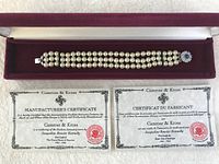 Triple strand faux pearl bracelet with blue sapphire-like clasp displayed in burgundy velvet box alongside two Certificates of Authenticity from Camrose & Kross.