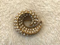 Close-up photo of the gold-tone swirl brooch encrusted with clear rhinestones against a cream fabric background